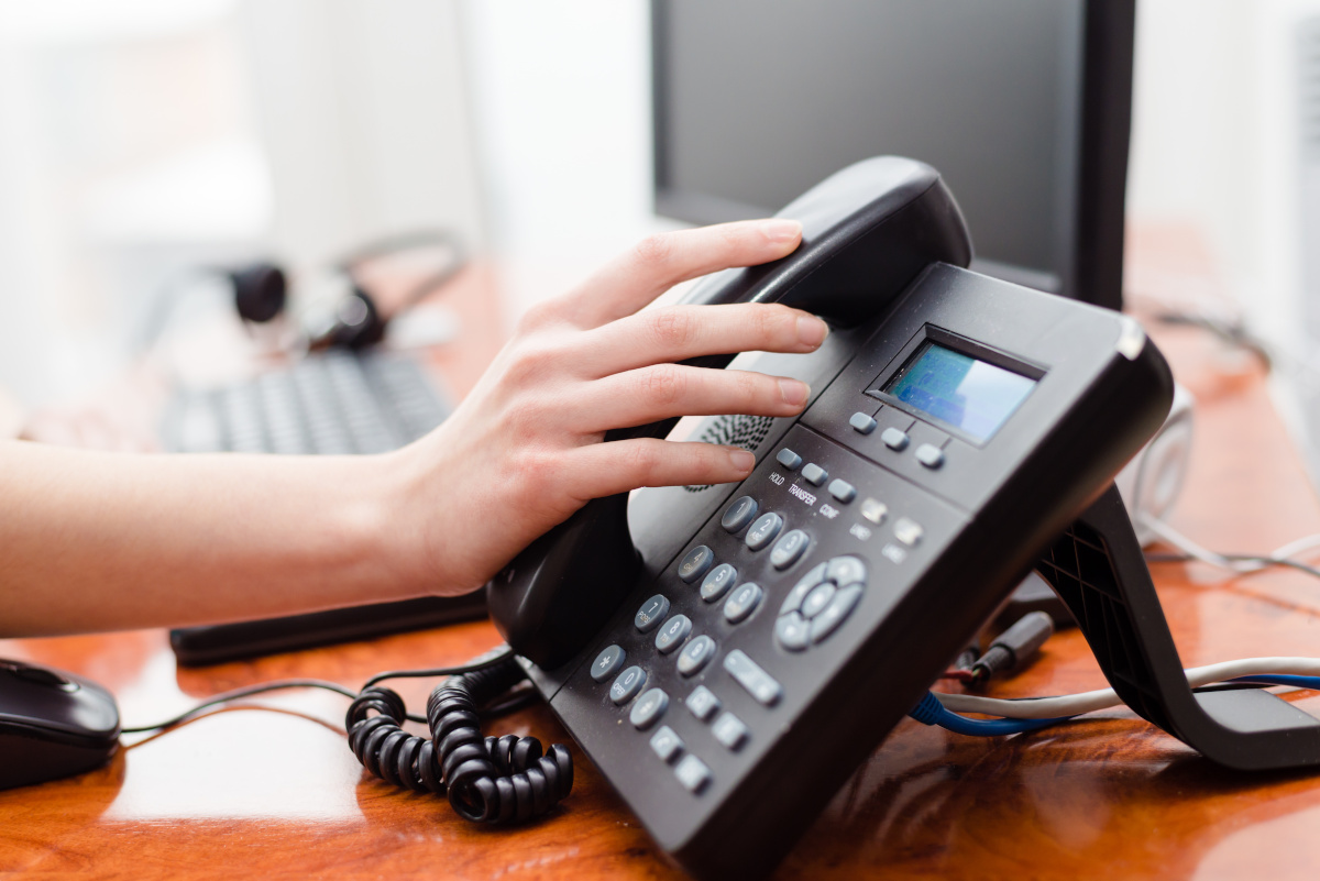 A hand resting on a phone receiver that is on its base, with a keyboard, monitor & headset blurred in the background