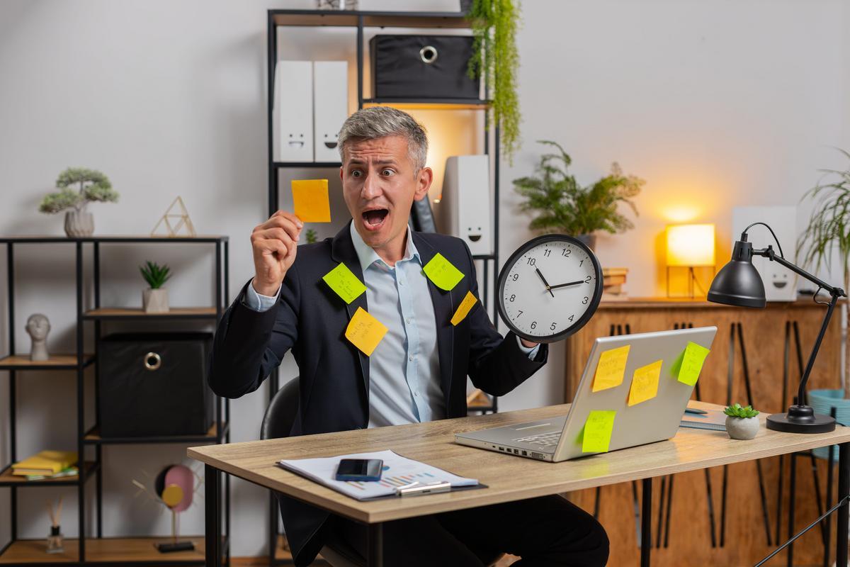 Stressed business owner at a home office desk holding a clock and sticky notes